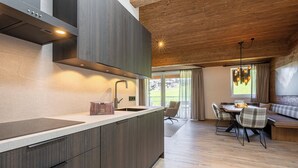 Ett modernt kök med en inbyggd köksö, en matplats med ett träbord och stolar samt utsikt över utomhusmiljön genom stora fönster.