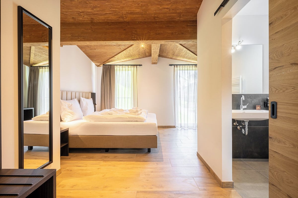 A bedroom with a wooden ceiling, a large bed, a bedside table, and a bathroom with a sink and mirror.