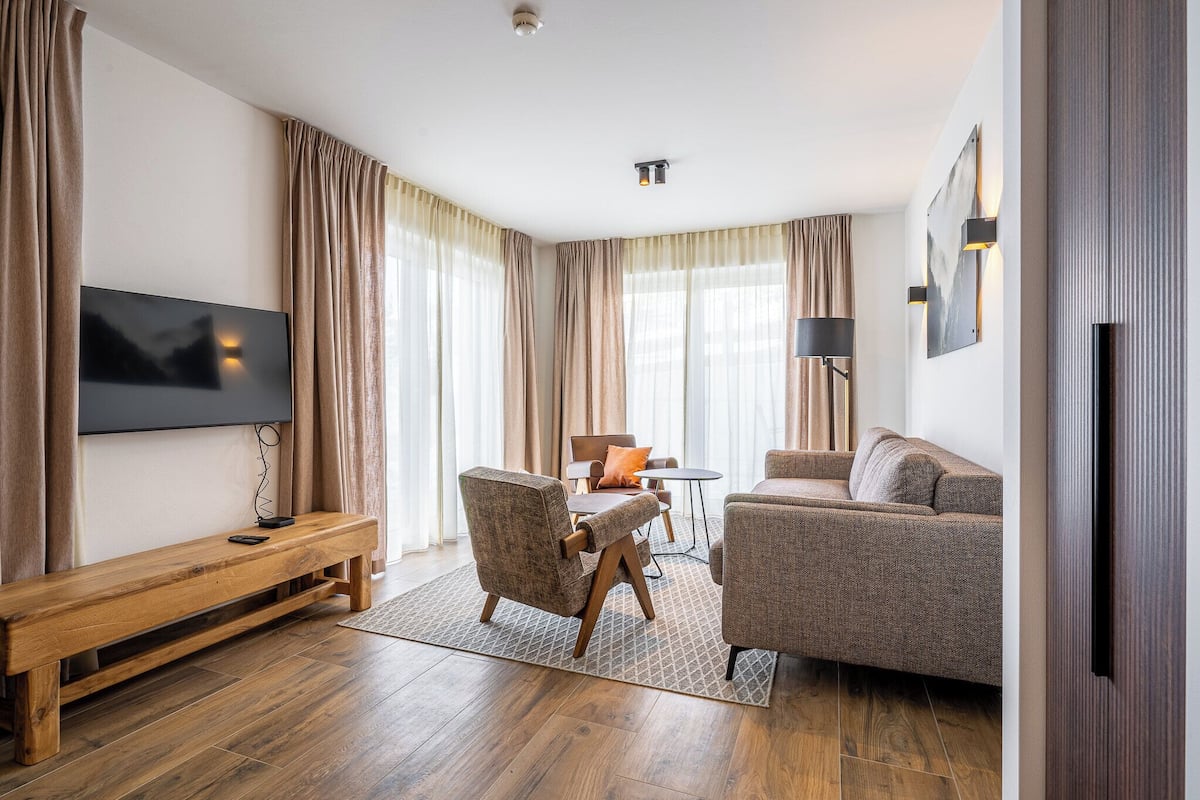 A modern living room with a flat-screen TV, a wooden bench, two armchairs, a coffee table, and a sofa.