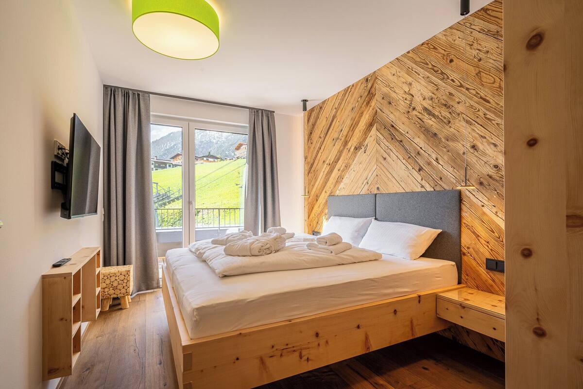 A bedroom with a wooden bed, a TV, a small wooden stool, and a window with curtains.
