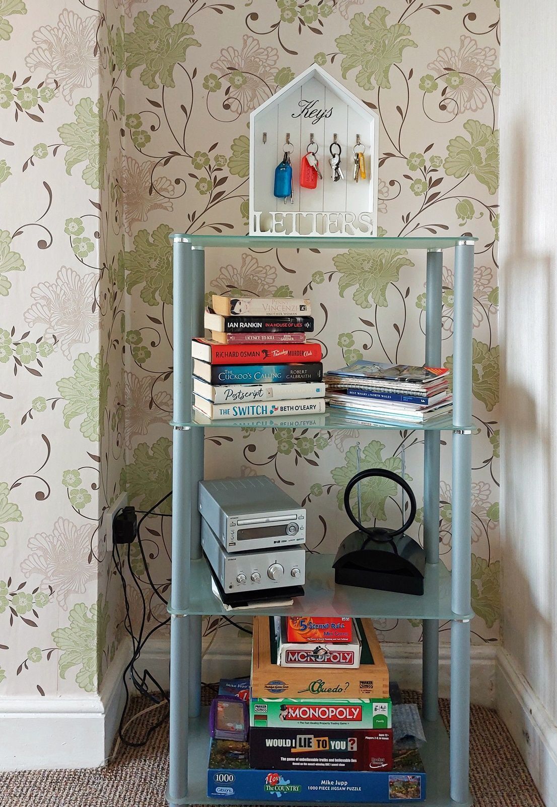 A bookshelf with various books and board games, a small house-shaped key holder, and a decorative wall with floral wallpaper.