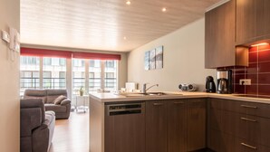 A modern kitchen with wooden cabinets, a dishwasher, and a coffee maker. There is a dining area with a sofa and a window with a red curtain. - Apartment With Lateral sea View in Ostend (Ostend)