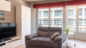 A modern living room with a grey sofa, a flat-screen TV, a small side table with a plant, and large windows with a red valance. - Apartment With Lateral sea View in Ostend (Ostend)
