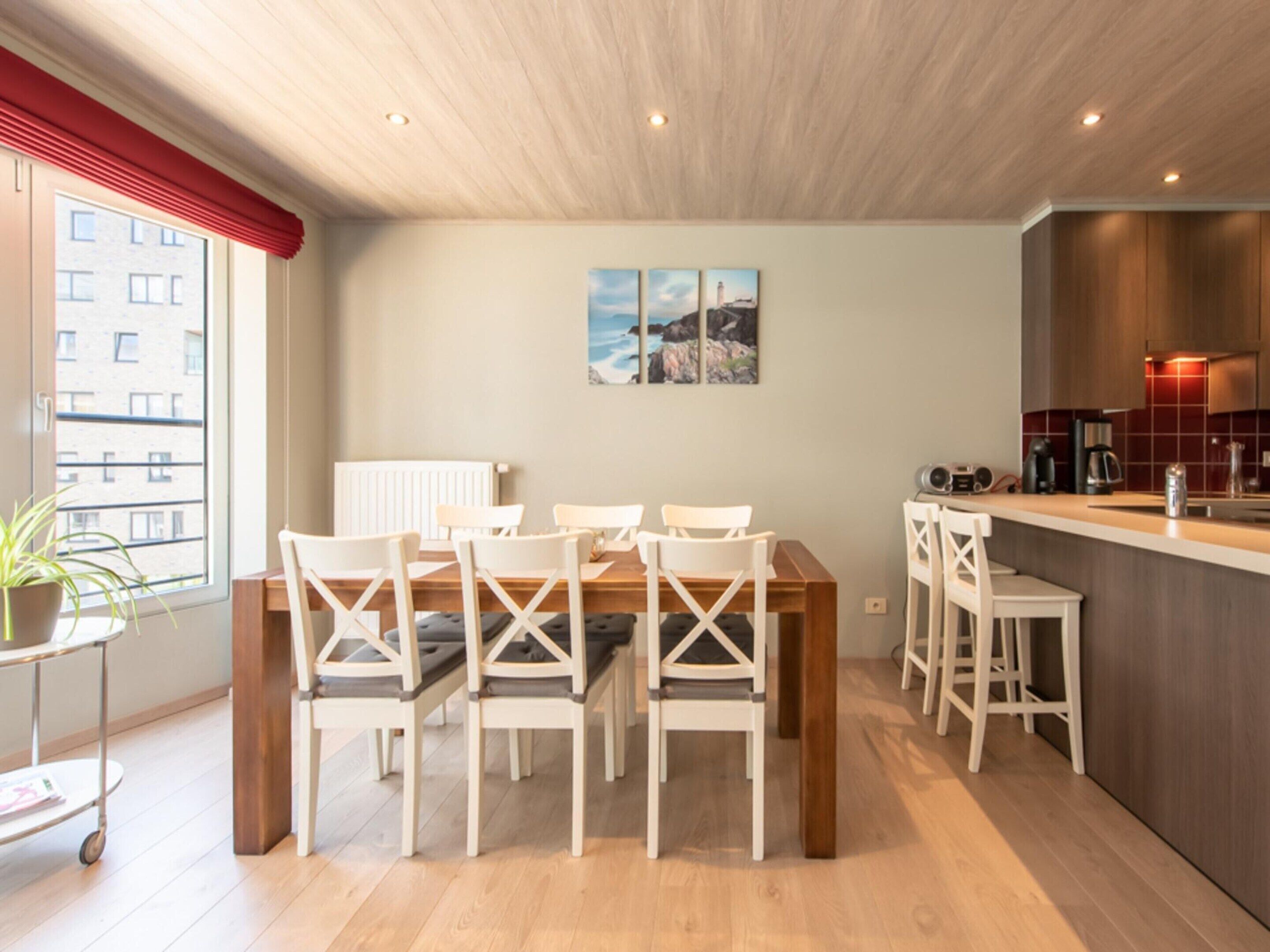 A modern kitchen with a wooden countertop and white chairs. There is a dining table with a white tablecloth and a potted plant on the side.