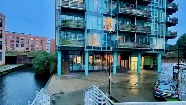 Een modern gebouw met meerdere balkons, een gracht ervoor en een auto in de buurt geparkeerd.