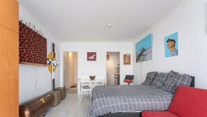 A compact hotel room with a bed, a red sofa, a wooden table with a bowl and candle, a small desk with a chair, and a wall with patterned wallpaper and artwork. - Studio in Blankenberge Near Beach & Pier (Blankenberge)