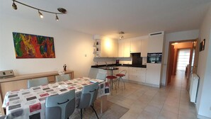A modern kitchen with white cabinets, a black countertop, and a built-in oven. There is a dining area with a table and chairs, and a colorful abstract painting on the wall. - Comfy City Stay Near Sea (Ostend)