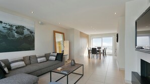 Ein modernes Wohnzimmer mit einem grauen Sofa, einem Couchtisch und einem Fernseher. Im Hintergrund ist eine Essecke mit Tisch und Stühlen zu sehen.