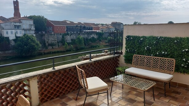 Ein Balkon mit Blick auf einen Fluss, eine Steinmauer und eine Reihe von Gebäuden im Hintergrund.