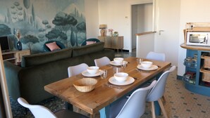 Ein Essbereich mit einem Holztisch, gedeckt für vier Personen, umgeben von weißen Stühlen. Hinter dem Tisch hängt ein großes Gemälde von Bäumen.