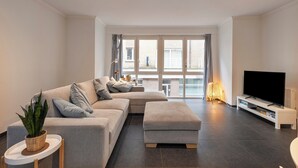 A modern living room with a grey sectional sofa, a white ottoman, a flat-screen TV on a white stand, and a large window with curtains.