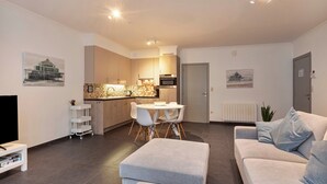 A modern living room with a grey sofa, a dining area with a white table and chairs, and a kitchen with wooden cabinets and a microwave.