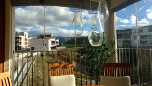 Un balcon avec deux chaises en bois, un vase en verre et une vue sur des immeubles et des montagnes.