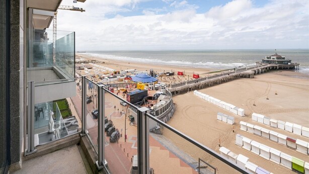 Vue sur la plage depuis un balcon doté d’une balustrade en verre, avec une jetée et des parasols de plage en arrière-plan.