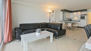 A modern living room with a black leather sofa, a white coffee table, and a dining area with a small kitchen in the background. - Luxbeach Apartment in Blankenberge (Blankenberge)