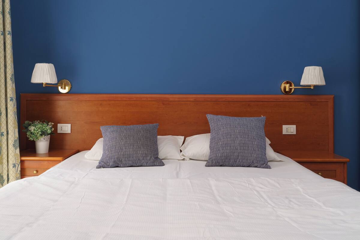 A neatly made bed with white linens and two patterned pillows, a wooden headboard, and two wall-mounted lamps with white shades.