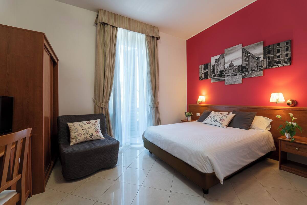 A hotel room with a bed, a chair, a TV, a window with curtains, and wall art.