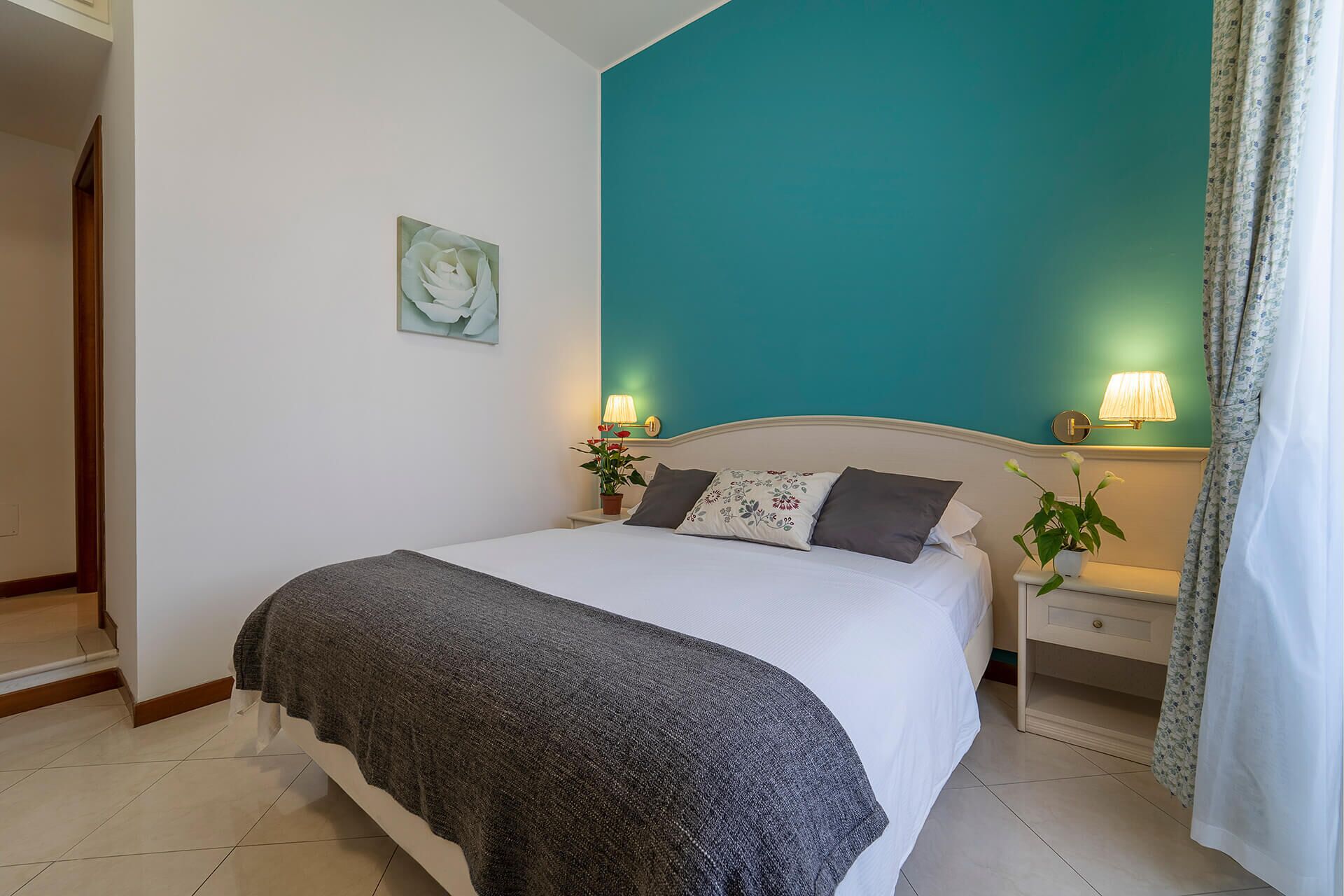 A bedroom with a large bed, two bedside tables with lamps, a potted plant, and a teal accent wall.