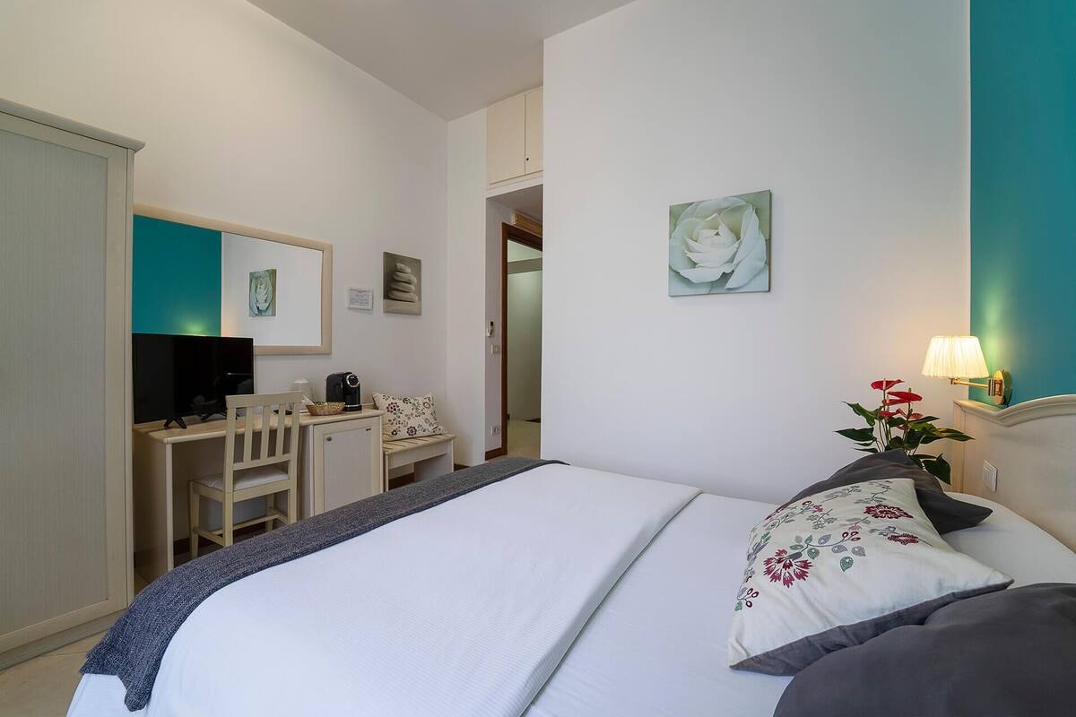 A hotel room with a bed, a desk with a computer, a chair, a lamp, and a plant.