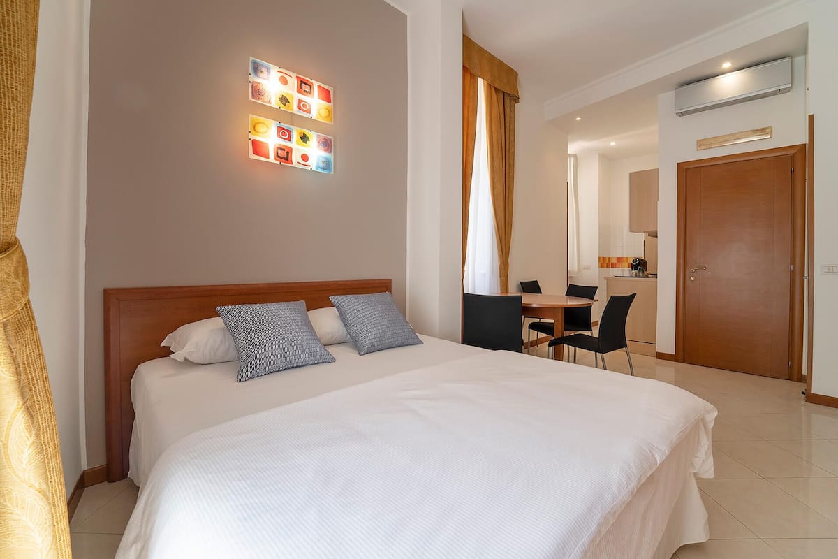 A hotel room with a large bed, two pillows, a desk, a chair, and a kitchenette.