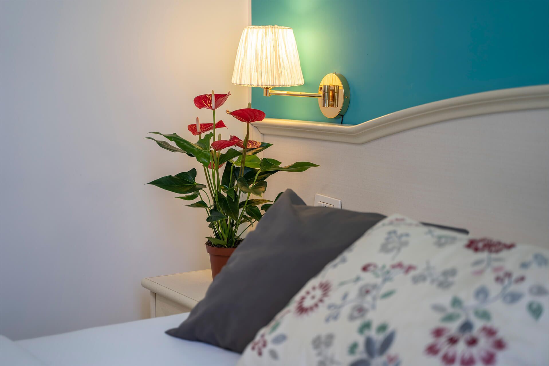 Un lit bien fait, agrémenté d’un coussin à motifs floraux et d’une plante en pot aux fleurs rouges.