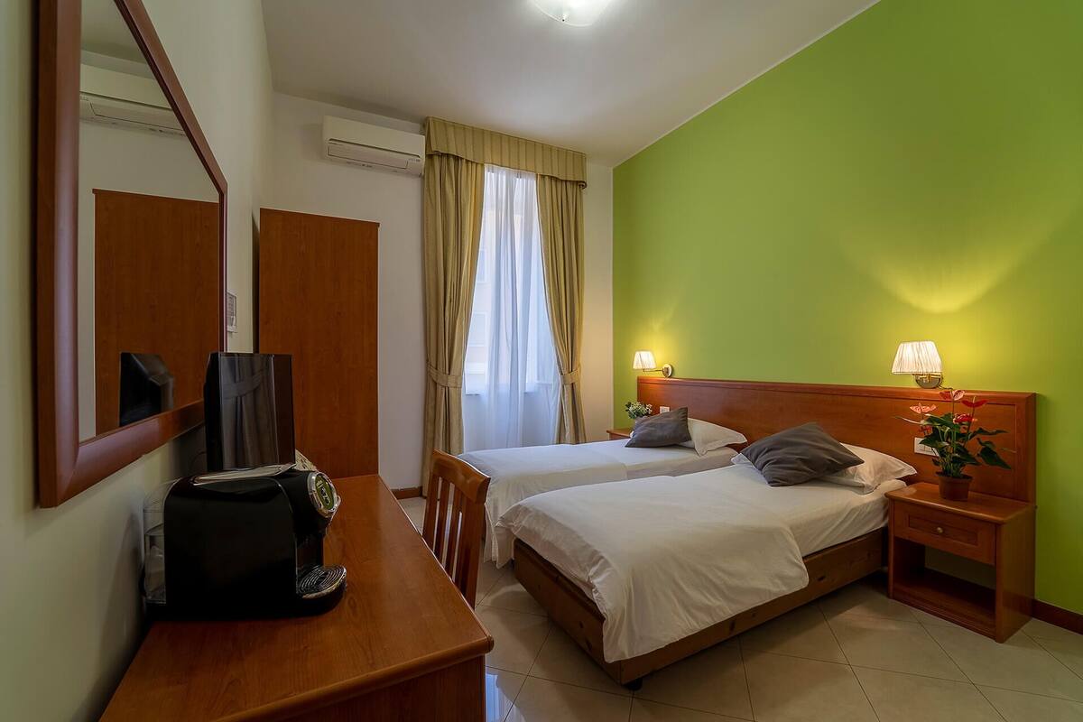 A hotel room with two beds, a desk with a coffee maker, a mirror, and a window with curtains.