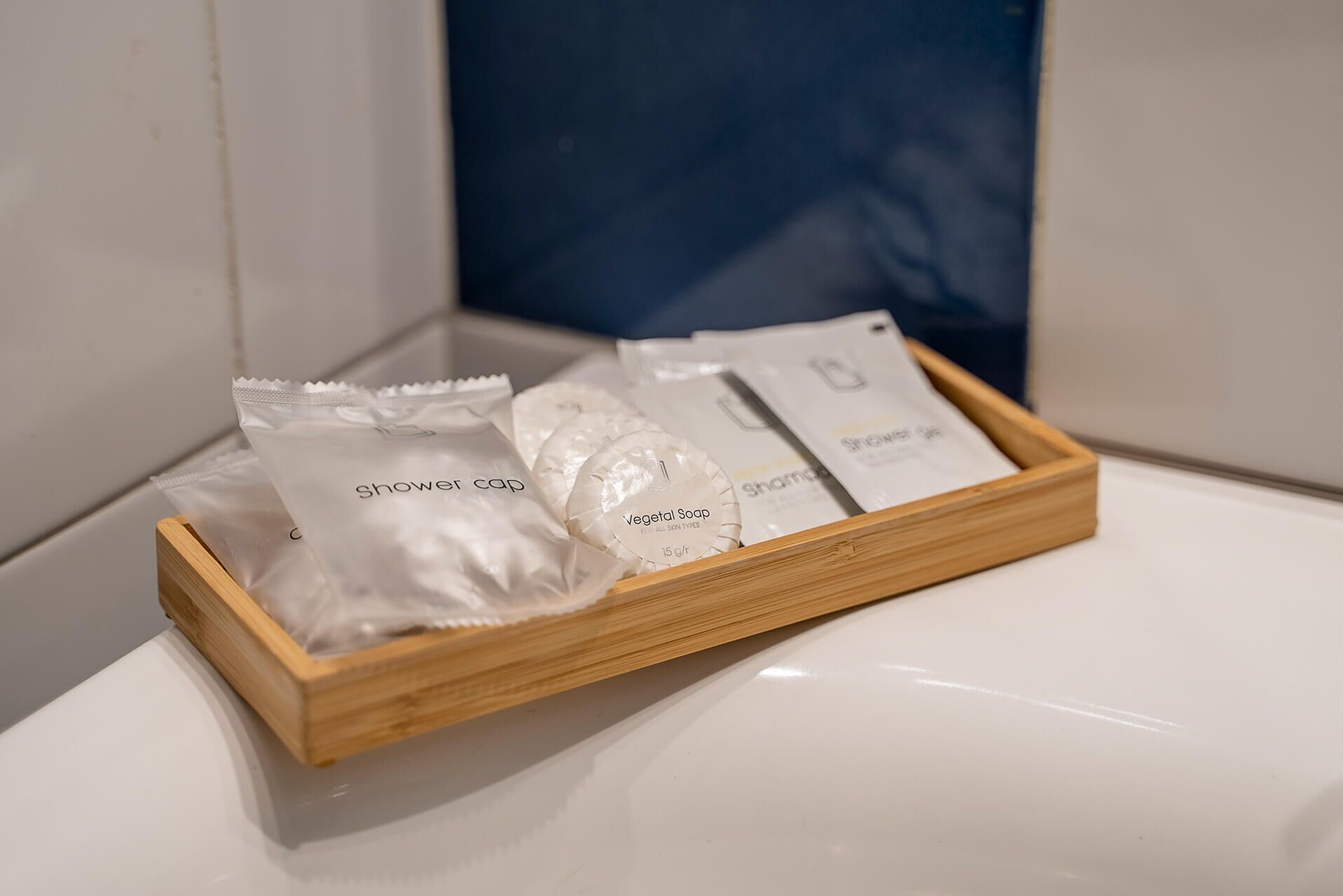 A wooden tray on a bathroom sink containing several toiletries including a shower cap, a bar of soap, and a packet of shampoo.