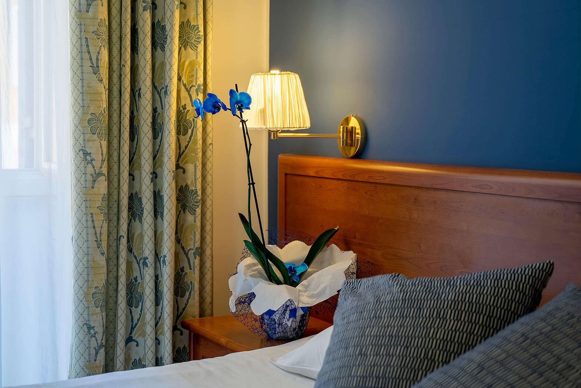 Ein Hotelzimmer mit Bett, Nachttisch mit Lampe, Vorhängen und einer Zierpflanze.