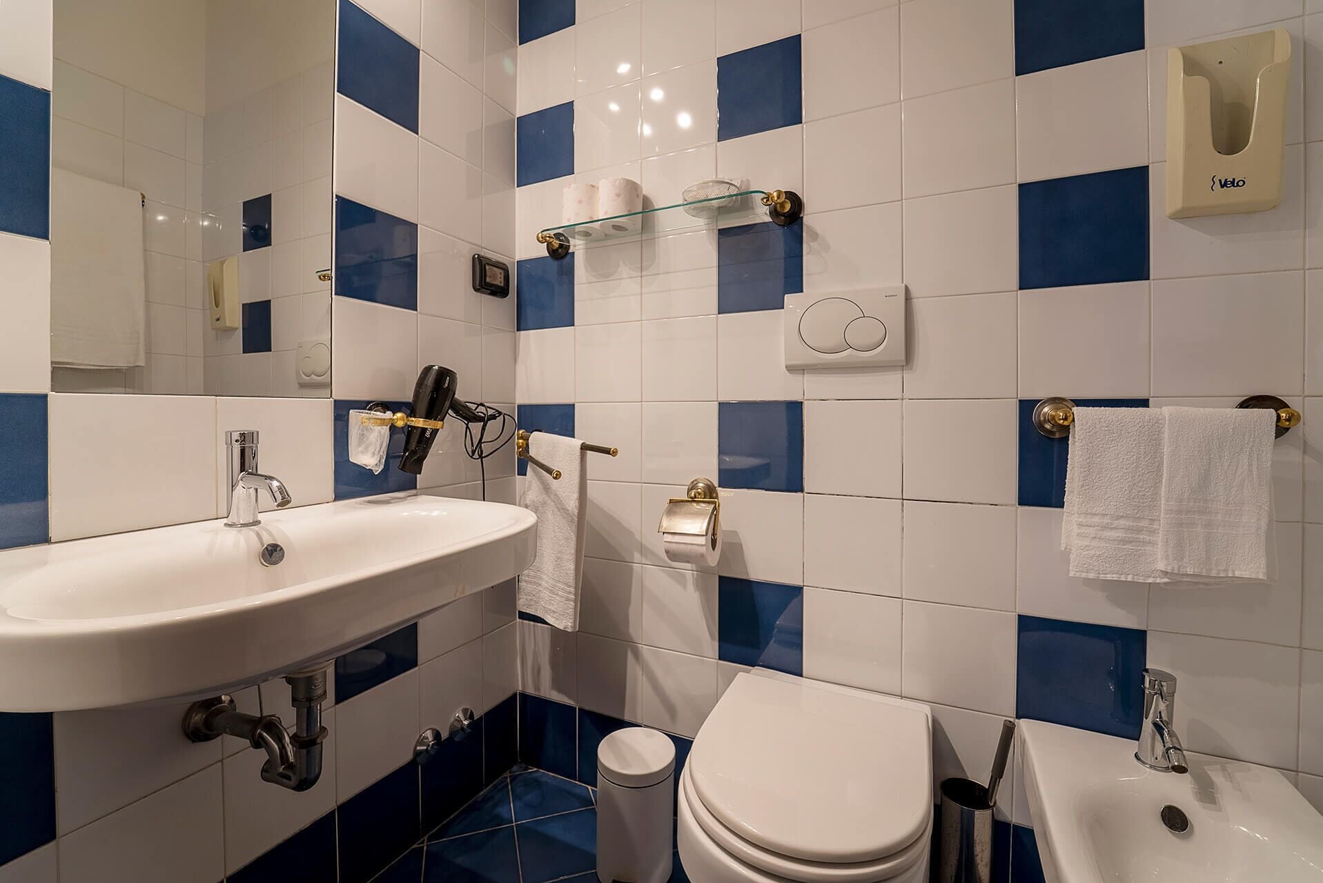 Un bagno con un lavandino bianco, un water e un bidet. Le pareti sono rivestite di piastrelle bianche e azzurre.