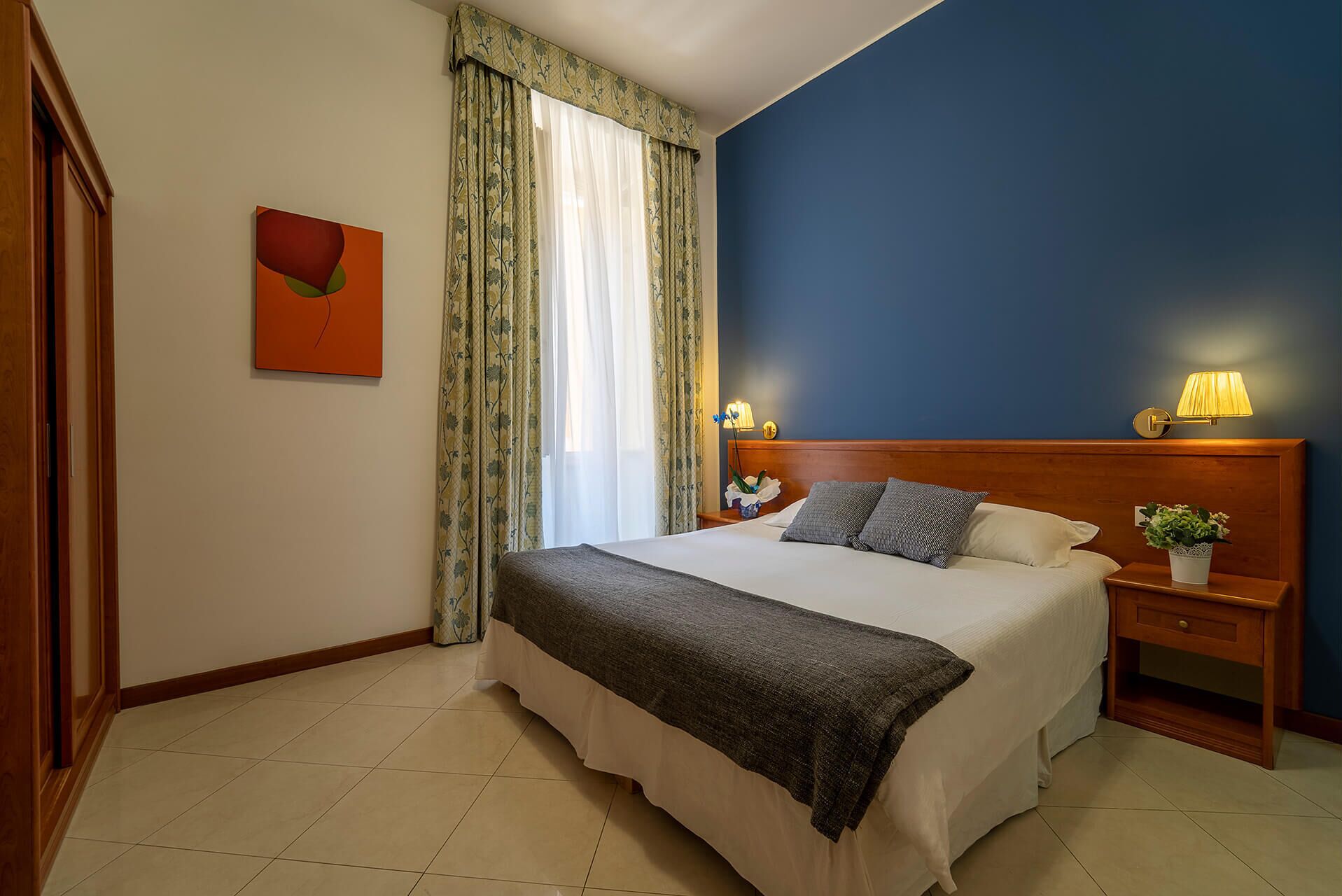 A hotel room with a bed, two bedside lamps, a nightstand, a potted plant, and a painting on the wall.
