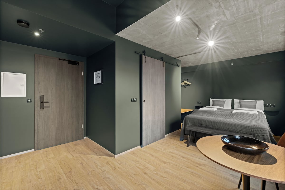 A modern hotel room with a wooden floor, a bed with grey bedding, a bedside table with a bowl, and a ceiling light fixture.