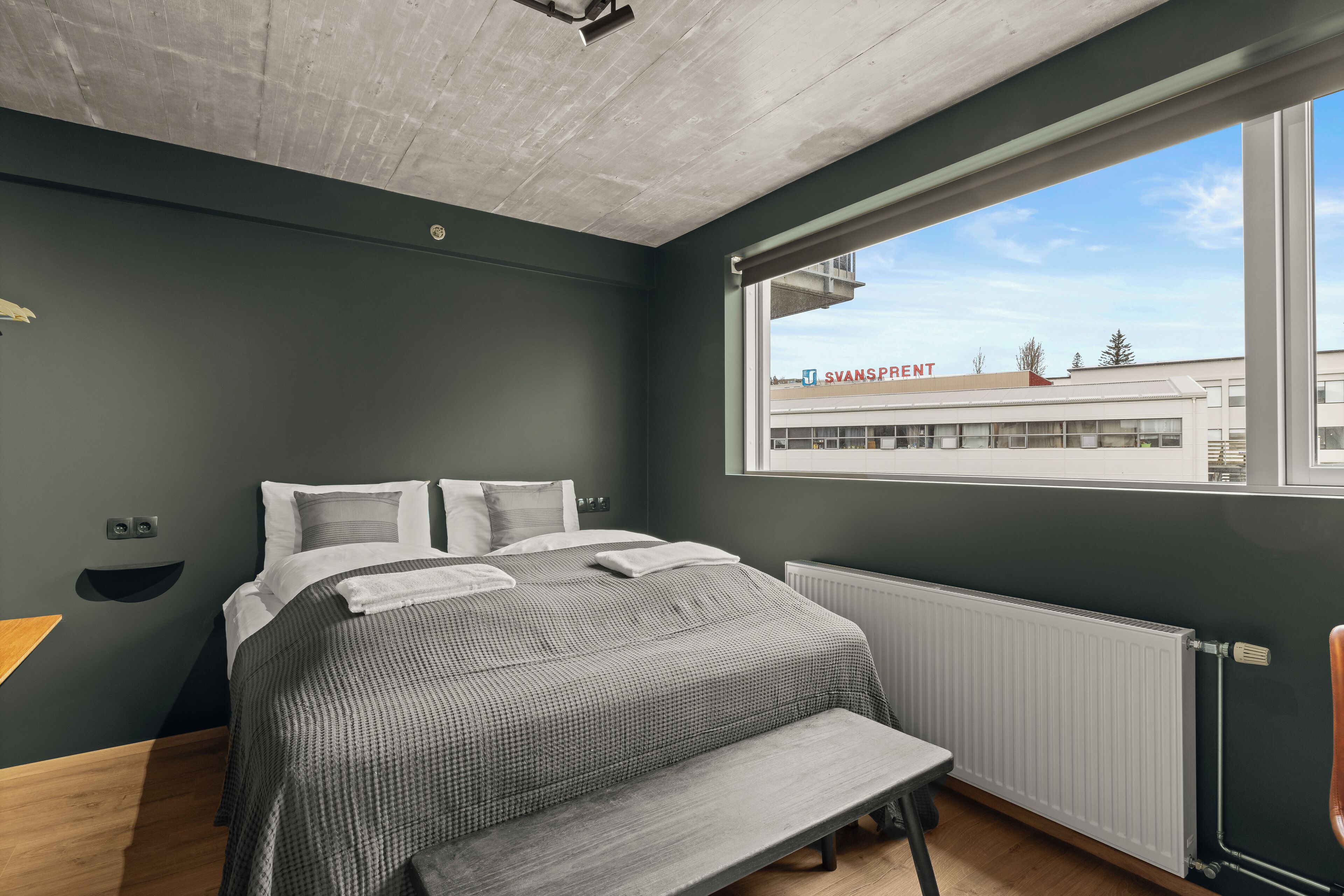 A modern bedroom with a large bed, a bench, a window with a view of a building, and a radiator.