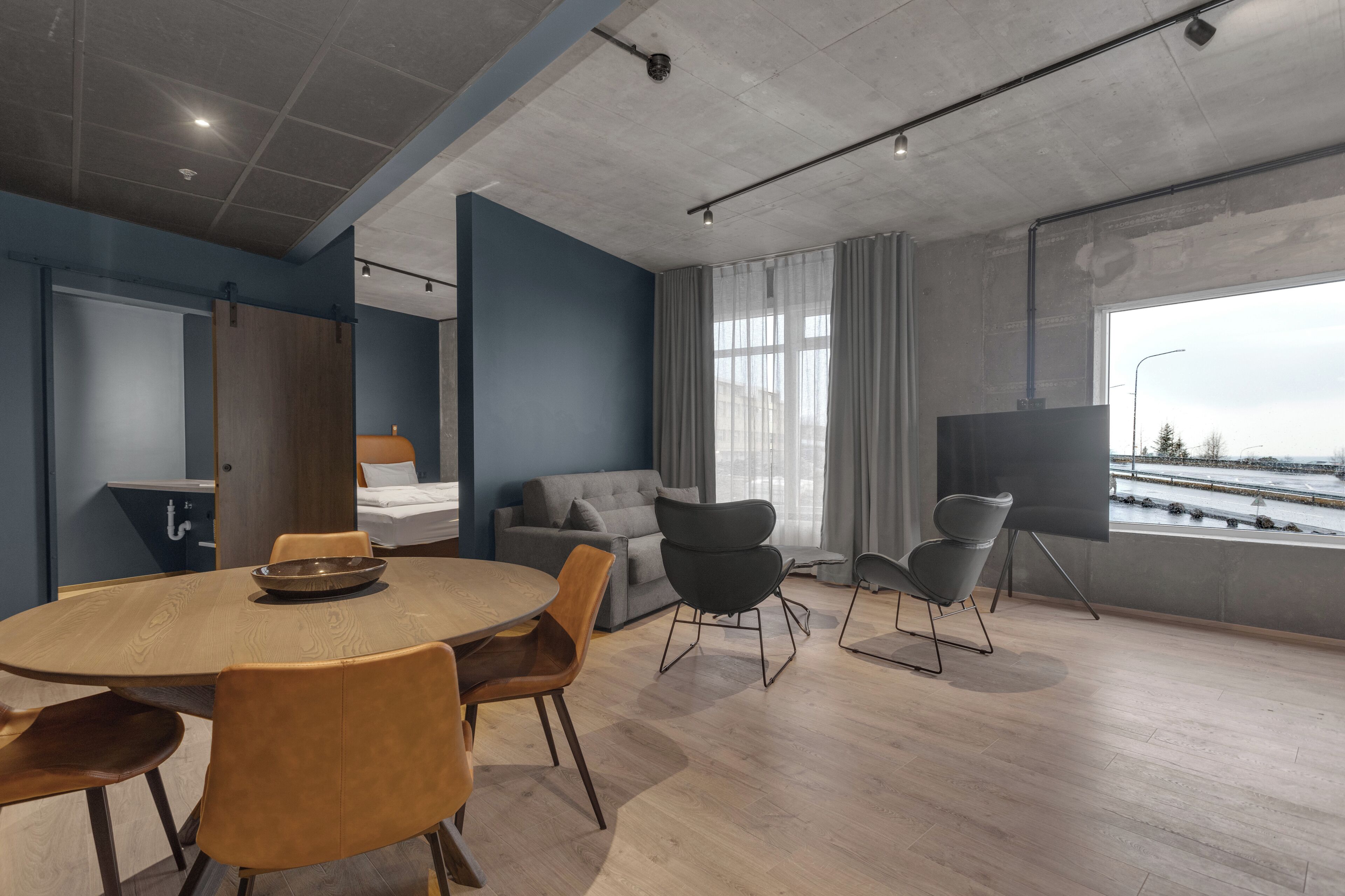 Een moderne kamer met een ronde houten tafel, verschillende stoelen, een bank, een televisie op een standaard en een groot raam met uitzicht op het water.