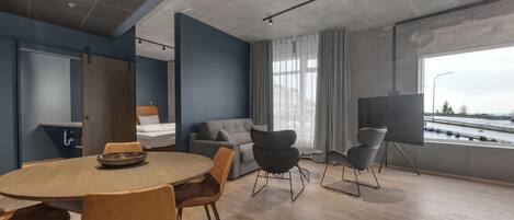Een moderne kamer met een ronde houten tafel, verschillende stoelen, een bank, een televisie op een standaard en een groot raam met uitzicht op het water.