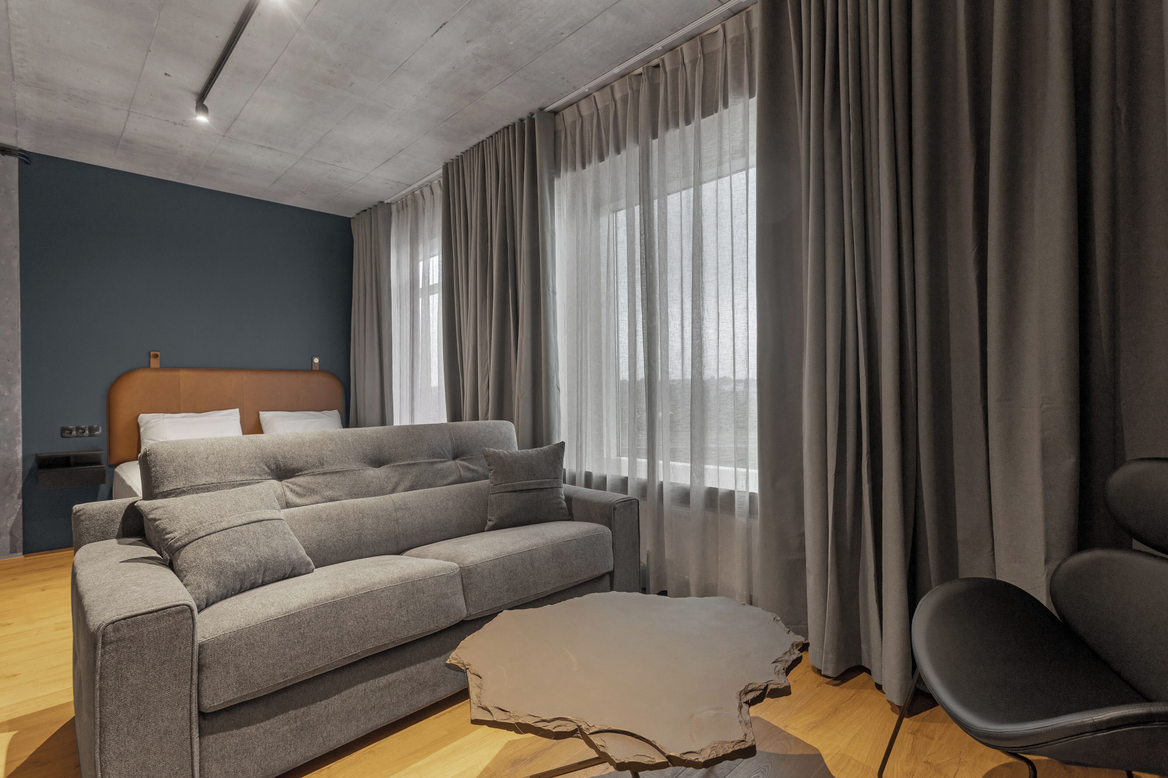 Una habitación de hotel moderna con un sofá gris, suelo de madera y un ventanal con cortinas grises.