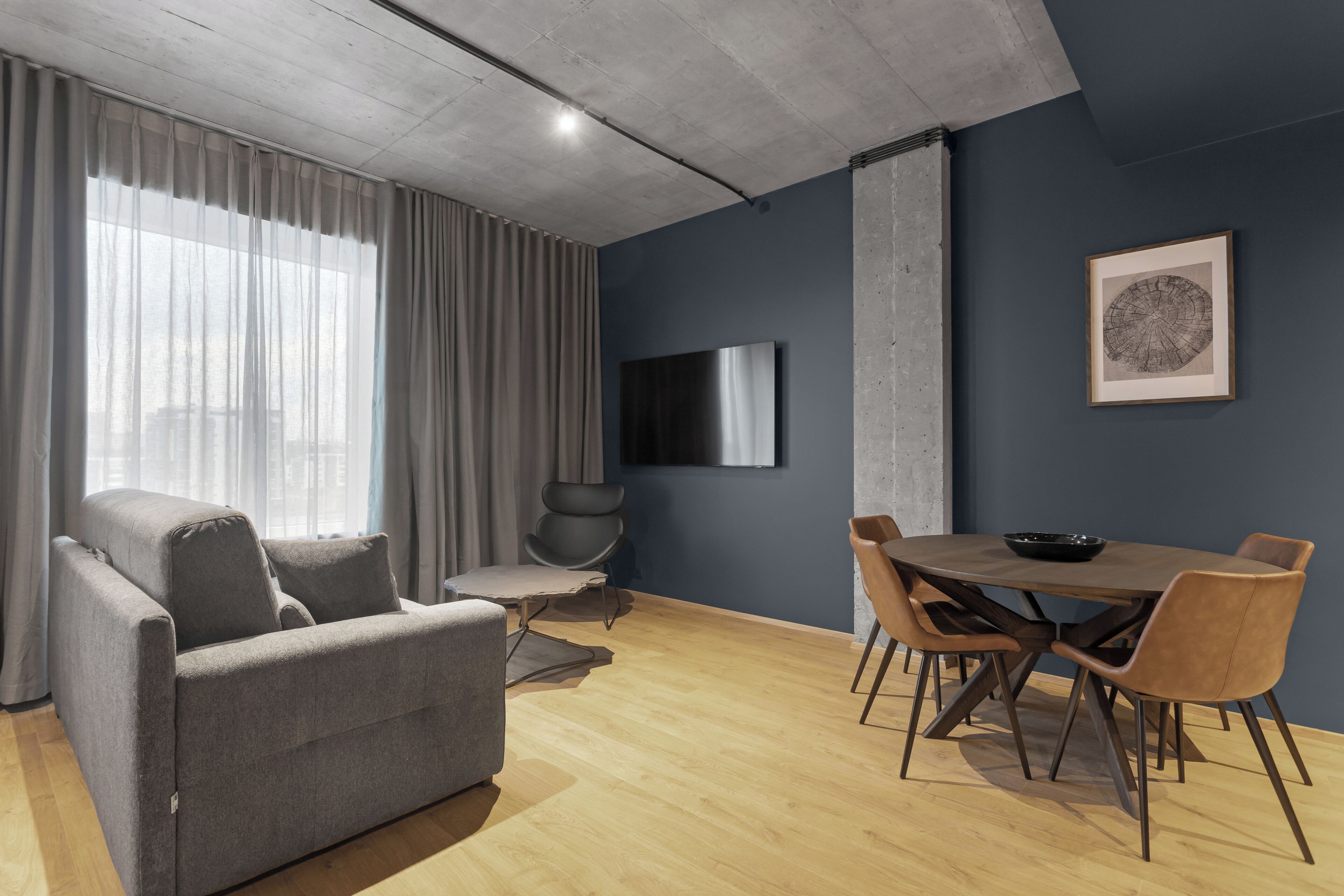 Una sala moderna con un sofá gris, una mesa de comedor con sillas, un televisor de pantalla plana montado en la pared y un gran ventanal con cortinas traslúcidas.