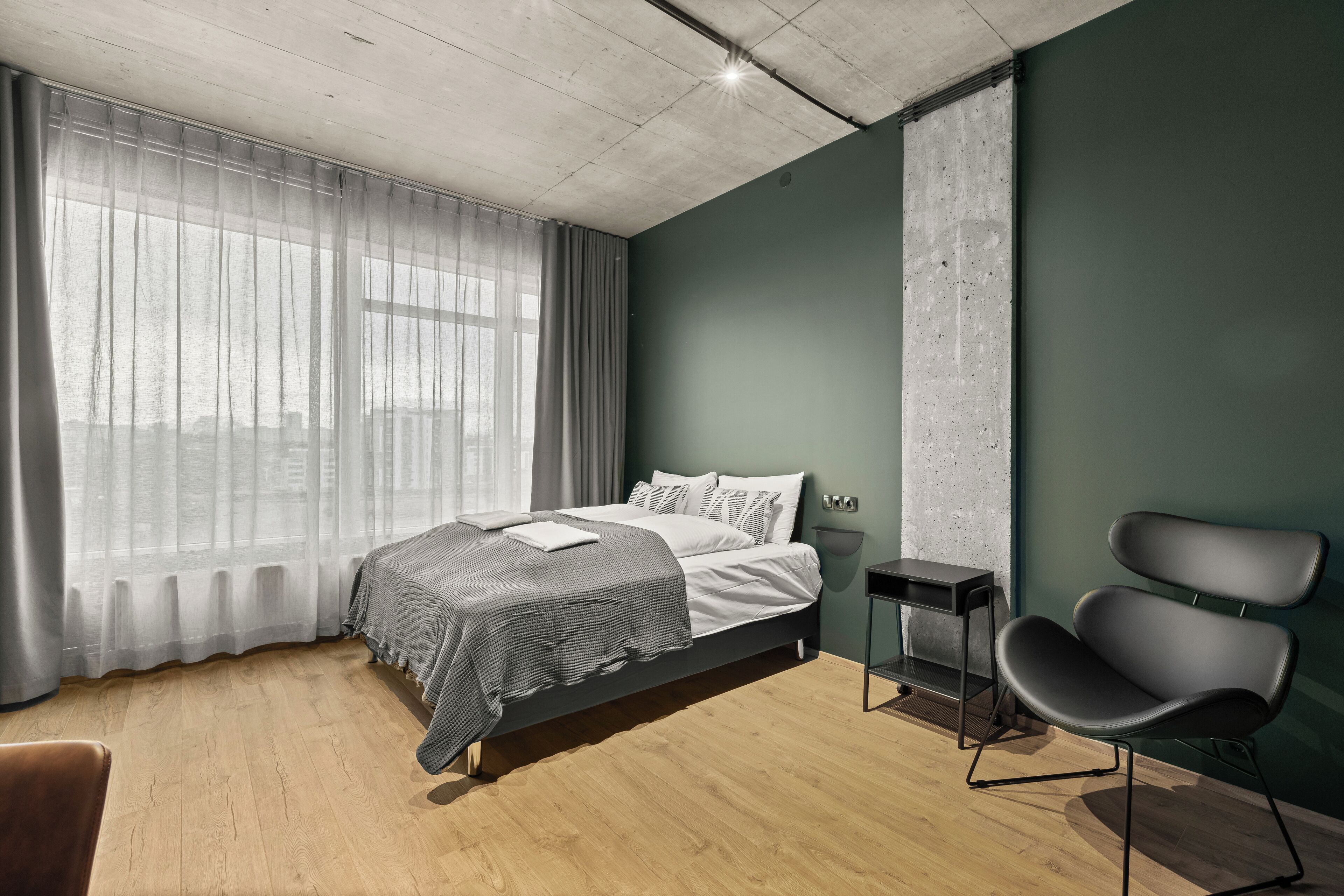 A modern bedroom with a bed, a chair, a nightstand, and a large window with sheer curtains.