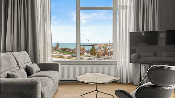 A modern living room with a grey sofa, a small table, and a TV. A large window offers a view of the sea and a distant cityscape. - Aurora Nooks (Kópavogur)