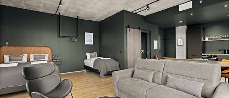 A modern hotel room with a large bed, a sofa, a chair, and a dining area with a kitchen in the background.