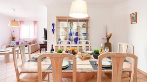 A dining area with a table set for four, surrounded by wicker chairs, a glass display cabinet with blue items, and a hanging lamp above. - Villa Azzurra (Albufeira)