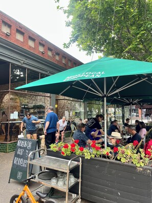 An outdoor seating area with a green umbrella, a menu board, and a cart with jars.