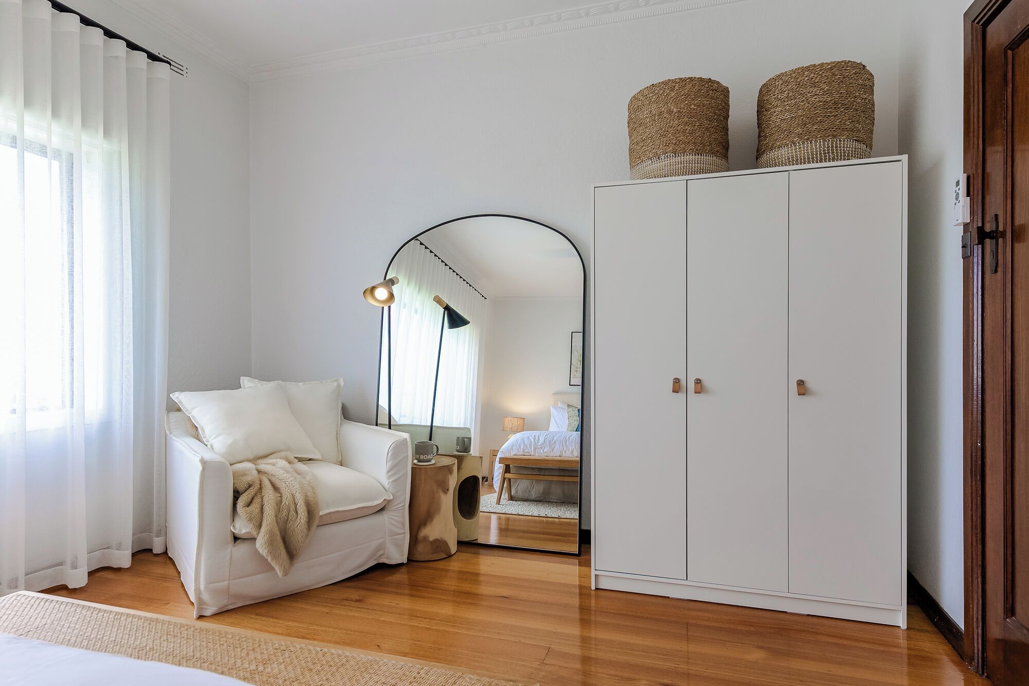 A bedroom with a white wardrobe, a white armchair, a small bedside table, and a mirror reflecting the room.