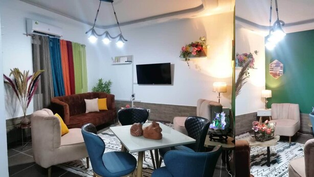 Un salon avec un canapé marron, des fauteuils bleus, une table blanche et un téléviseur à écran plat fixé au mur.