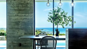 A modern dining area with a view of the ocean, a wooden table, a wicker chair, and a potted plant. - ILUVIO RESORT MOTOBU (Motobu)