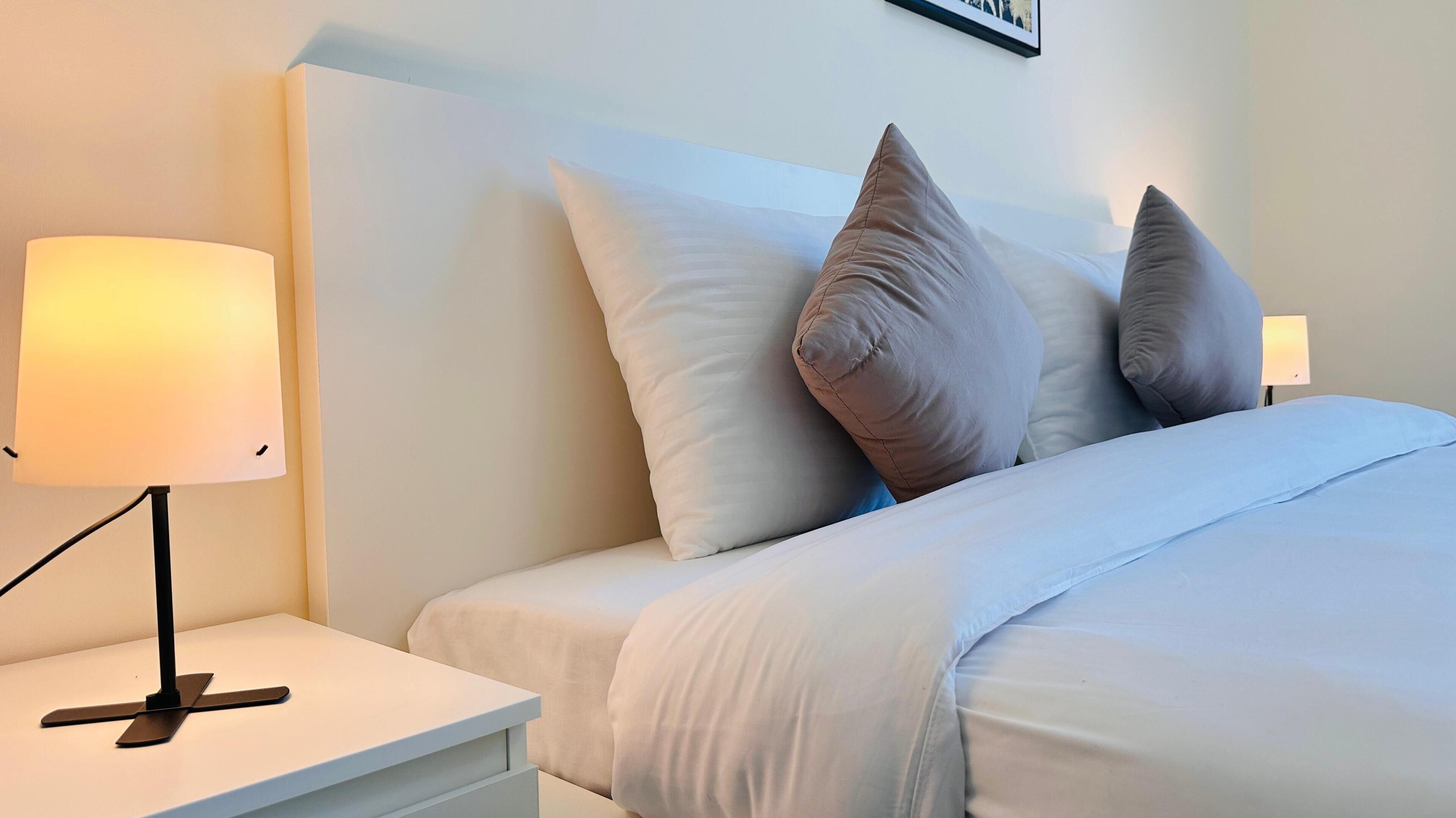 Una cama bien hecha con ropa de cama blanca y dos almohadas, una lámpara de noche y un cuadro en la pared.