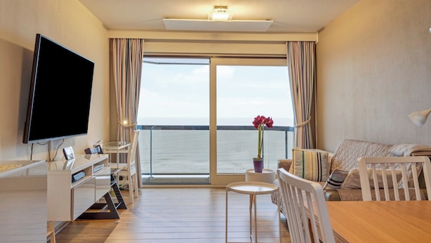 Une chambre moderne dotée d’une télévision à écran plat, d’une table à manger, d’un canapé et offrant une vue sur la mer grâce à de grandes fenêtres.