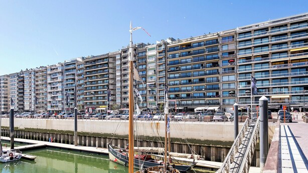 Una marina con barche ormeggiate, un edificio imponente con numerosi balconi, e un cielo sereno.