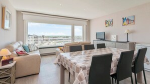 Una camera d'albergo moderna con una grande finestra, un'area pranzo con tavolo a scacchi, un divano e una televisione.