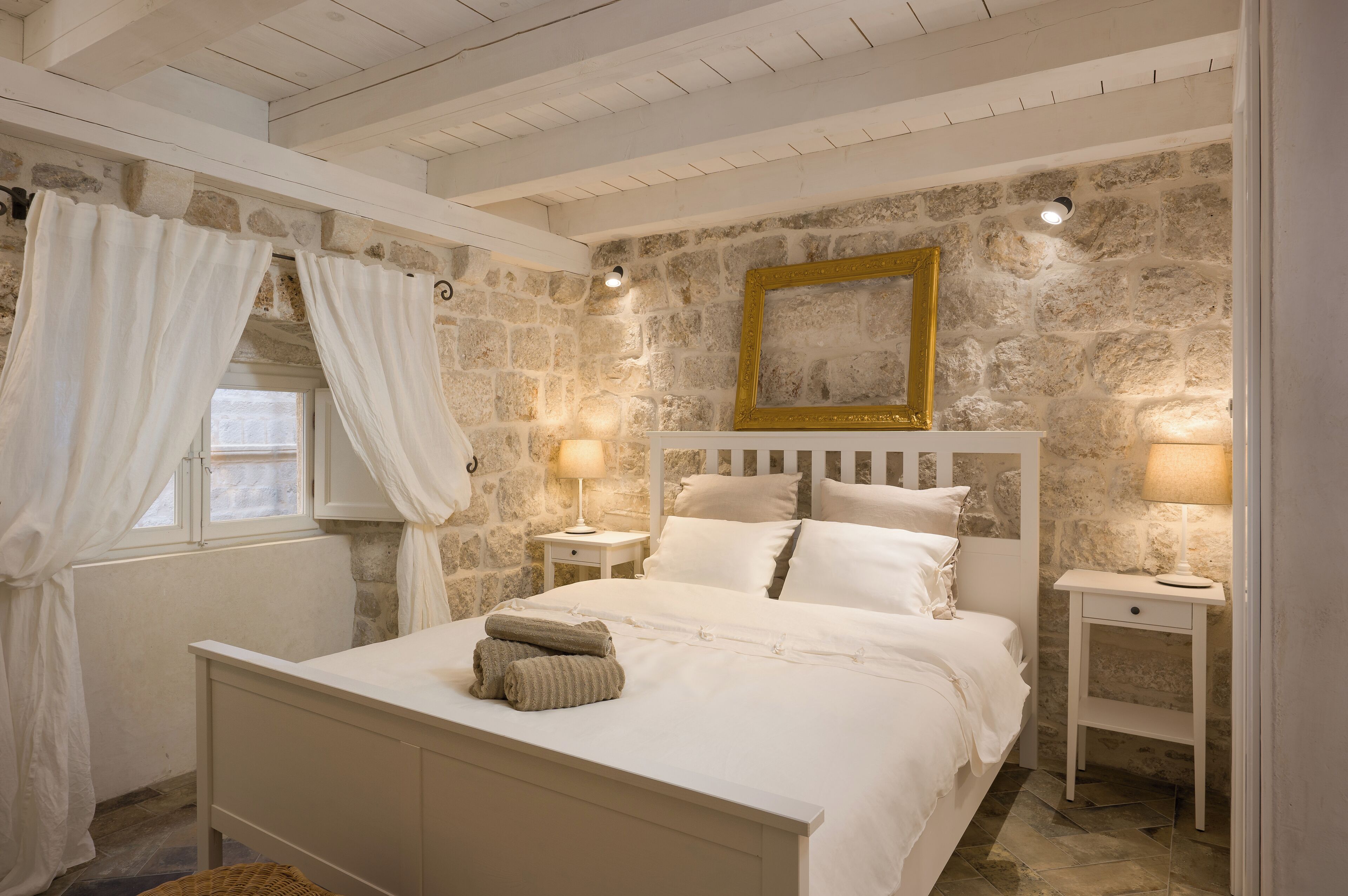 A bedroom with a stone wall, a bed with white bedding, two bedside tables with lamps, and a window with white curtains.