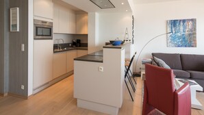 Una cucina moderna con mobili bianchi, forno integrato e microonde. È presente una piccola isola con una ciotola e una bilancia. Nell'area living ci sono una sedia rossa e un divano grigio.
